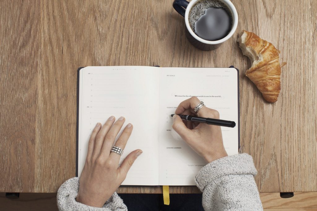Representative photograph of hands writing in an open notebook on a wooden table.